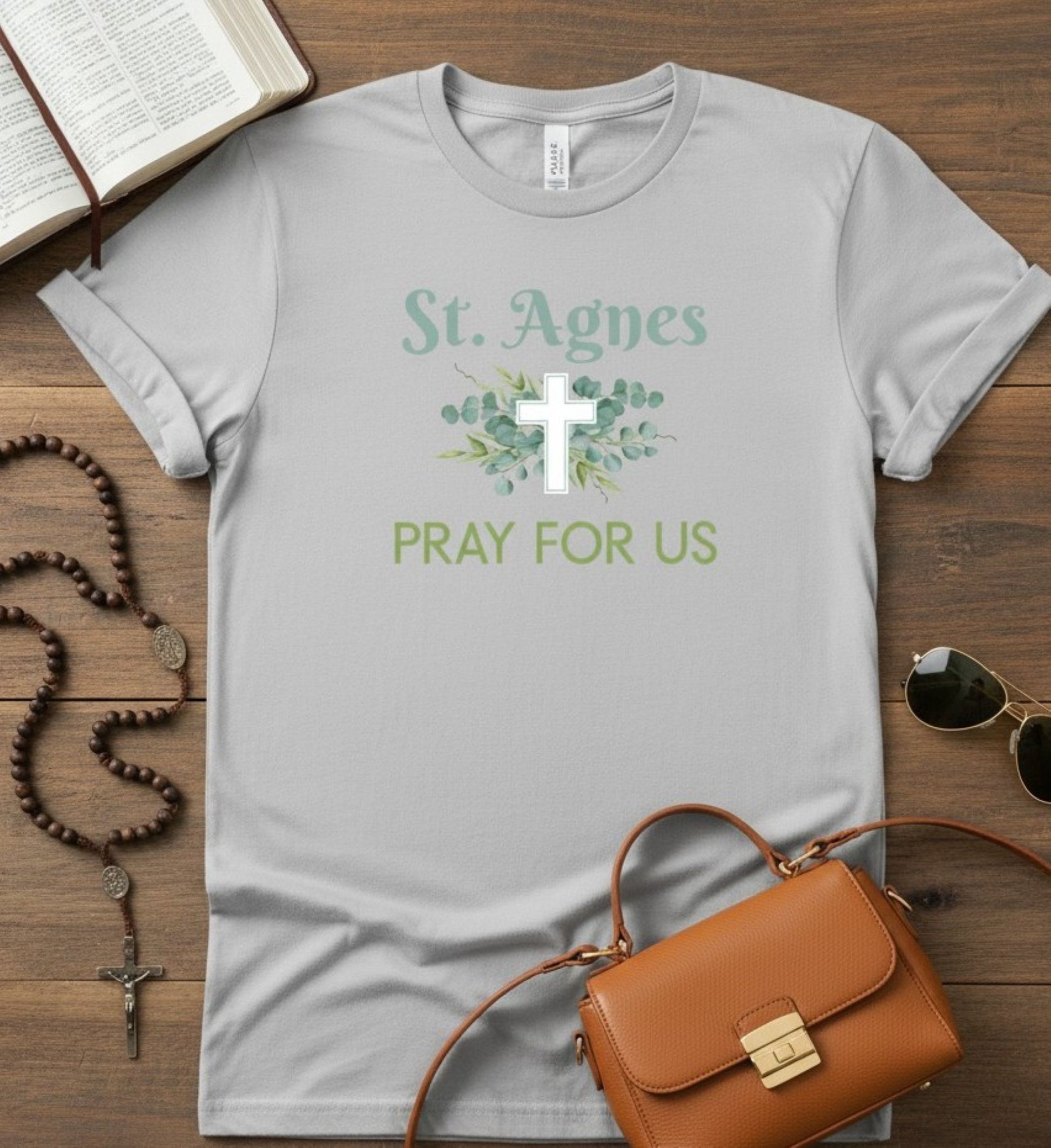 A light gray graphic t-shirt laid flat on a wooden surface, featuring a white cross, green eucalyptus leaves, and the text 'St. Agnes PRAY FOR US'.
