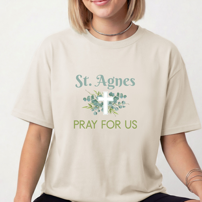 A woman wearing a beige crewneck t-shirt with a graphic print that reads 'St. Agnes PRAY FOR US' in sage green and white, featuring a white cross and eucalyptus leaves.