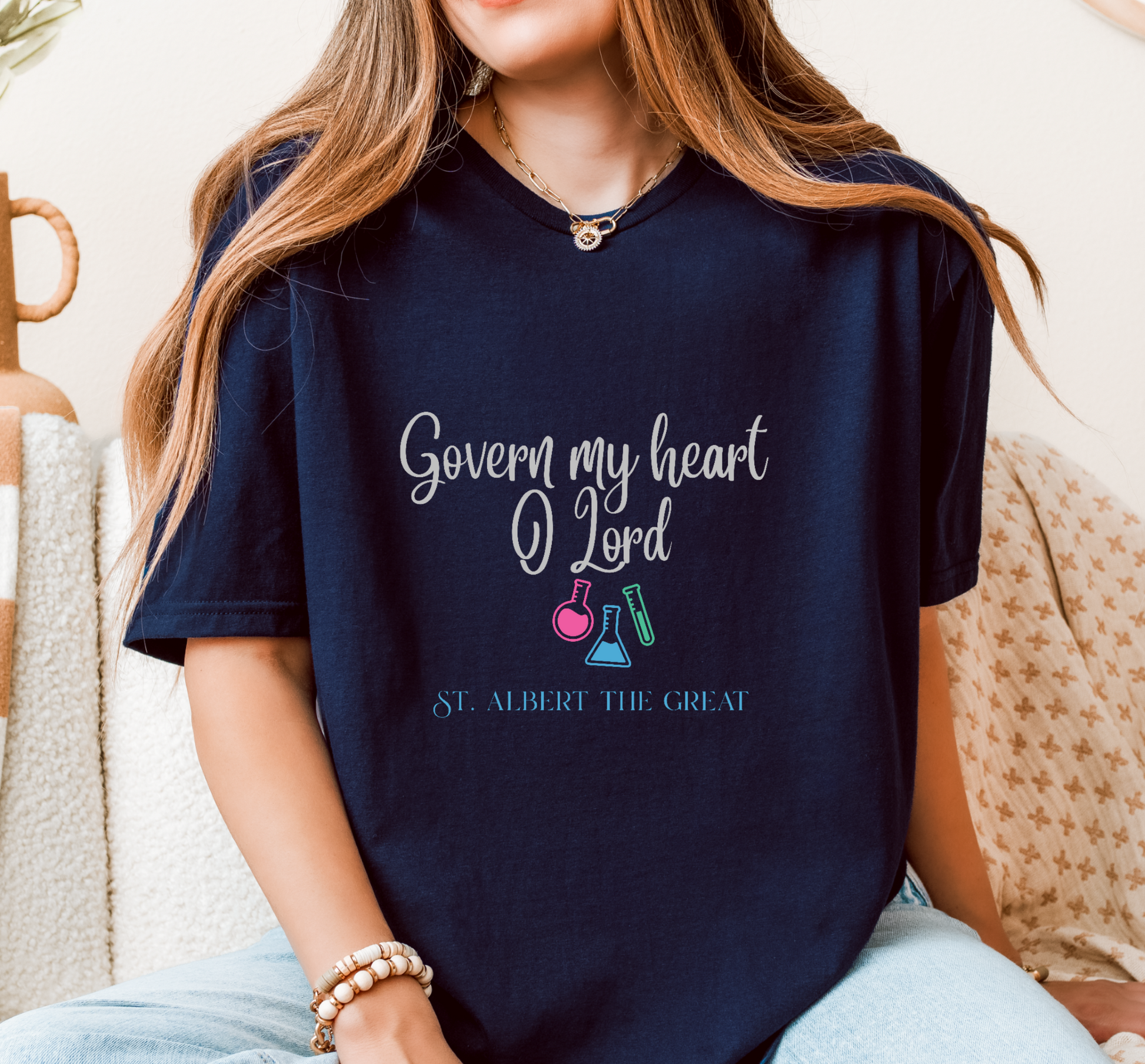 A woman wearing a navy blue graphic t-shirt with the text 'Govern my heart O Lord' and 'ST. ALBERT THE GREAT' along with colorful beaker illustrations, sitting on a patterned couch.