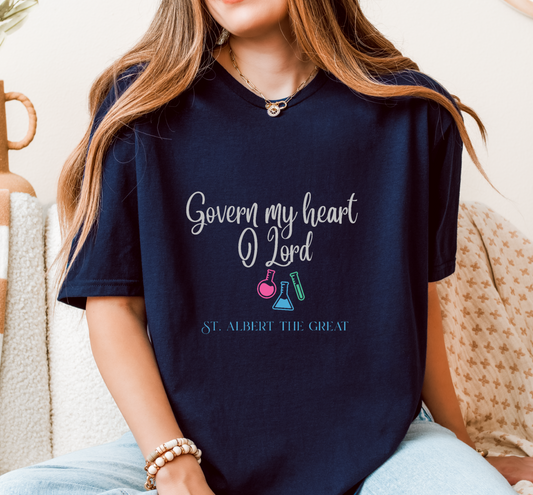 A woman wearing a navy blue graphic t-shirt with the text 'Govern my heart O Lord' and 'ST. ALBERT THE GREAT' along with colorful beaker illustrations, sitting on a patterned couch.