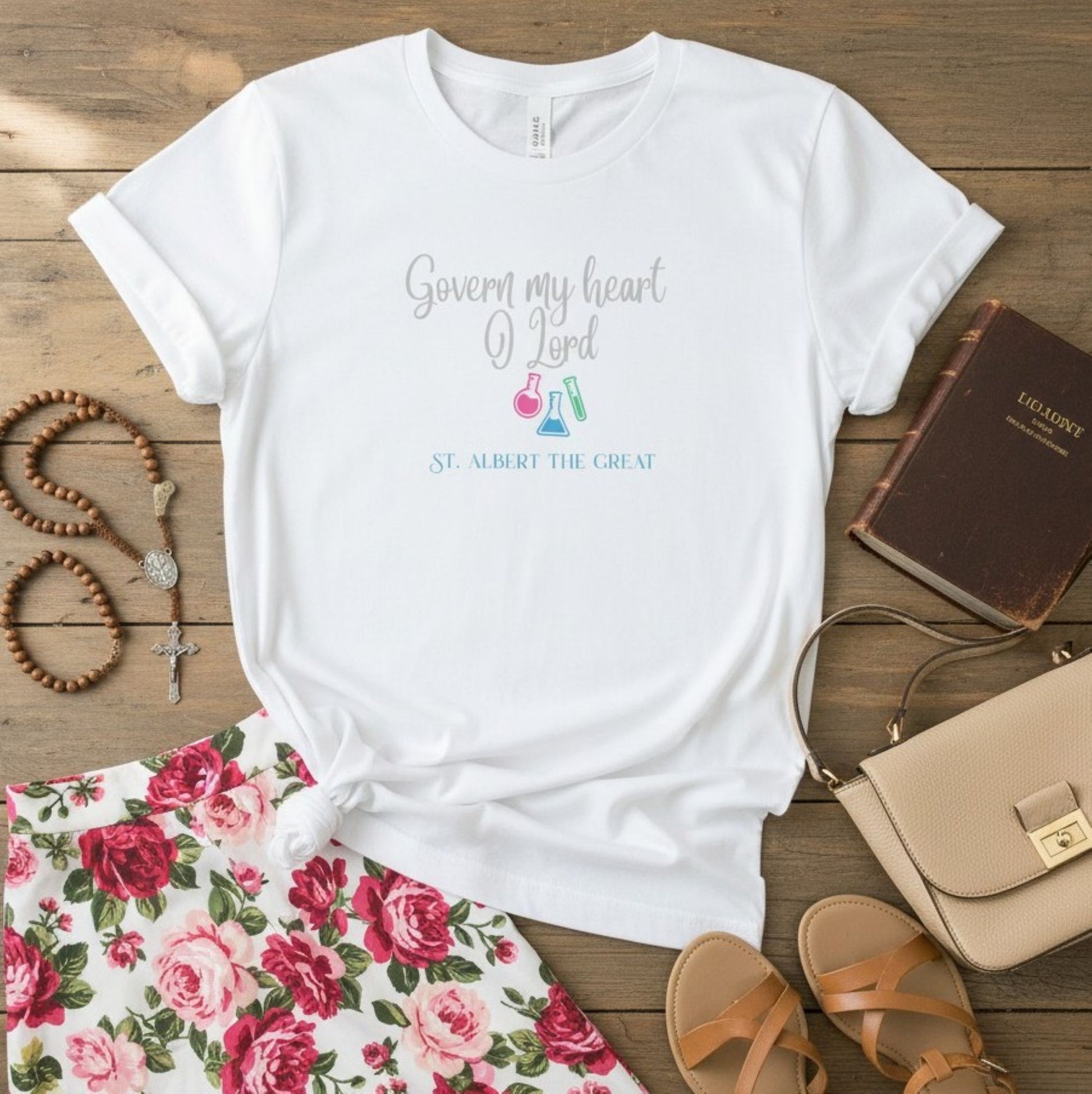 A white graphic T-shirt laid flat on a rustic wood surface, surrounded by styled props including a floral skirt, sandals, a handbag, a rosary, and a book. The shirt has a printed phrase reading 'Govern my heart O Lord' with small colorful flask illustrations and 'ST.