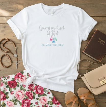 A white graphic T-shirt laid flat on a rustic wood surface, surrounded by styled props including a floral skirt, sandals, a handbag, a rosary, and a book. The shirt has a printed phrase reading 'Govern my heart O Lord' with small colorful flask illustrations and 'ST.