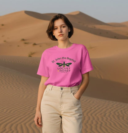 A woman stands in a desert sand dune setting wearing a bright pink graphic T-shirt and light beige pants. The shirt features a black and green bee graphic with the text 'St.
