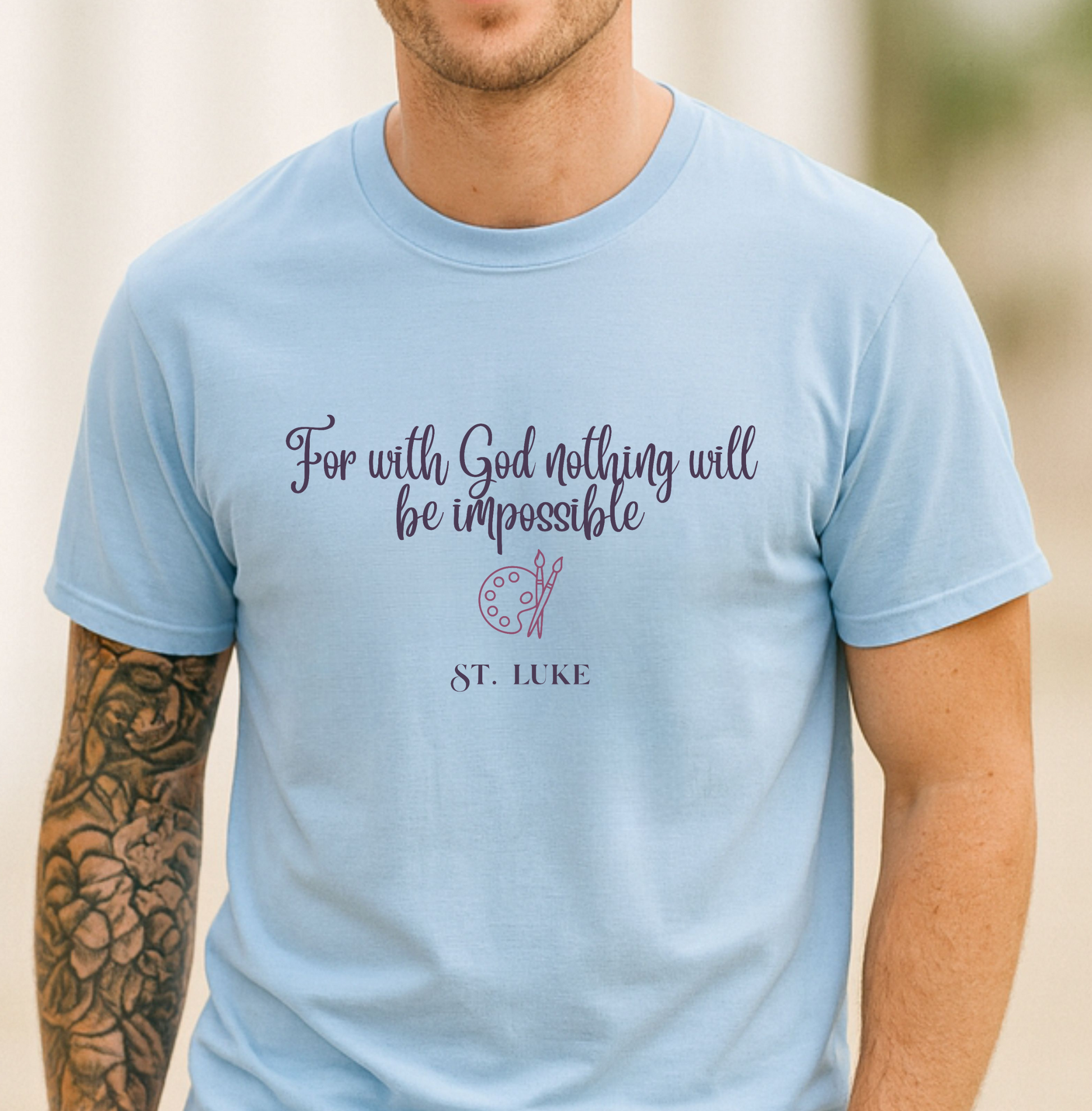 A man wearing a light blue crewneck t-shirt with a purple printed quote that reads 'For with God nothing will be impossible' and 'ST. LUKE' below it, set against a softly blurred outdoor background.