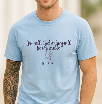 A man wearing a light blue crewneck t-shirt with a purple printed quote that reads 'For with God nothing will be impossible' and 'ST. LUKE' below it, set against a softly blurred outdoor background.