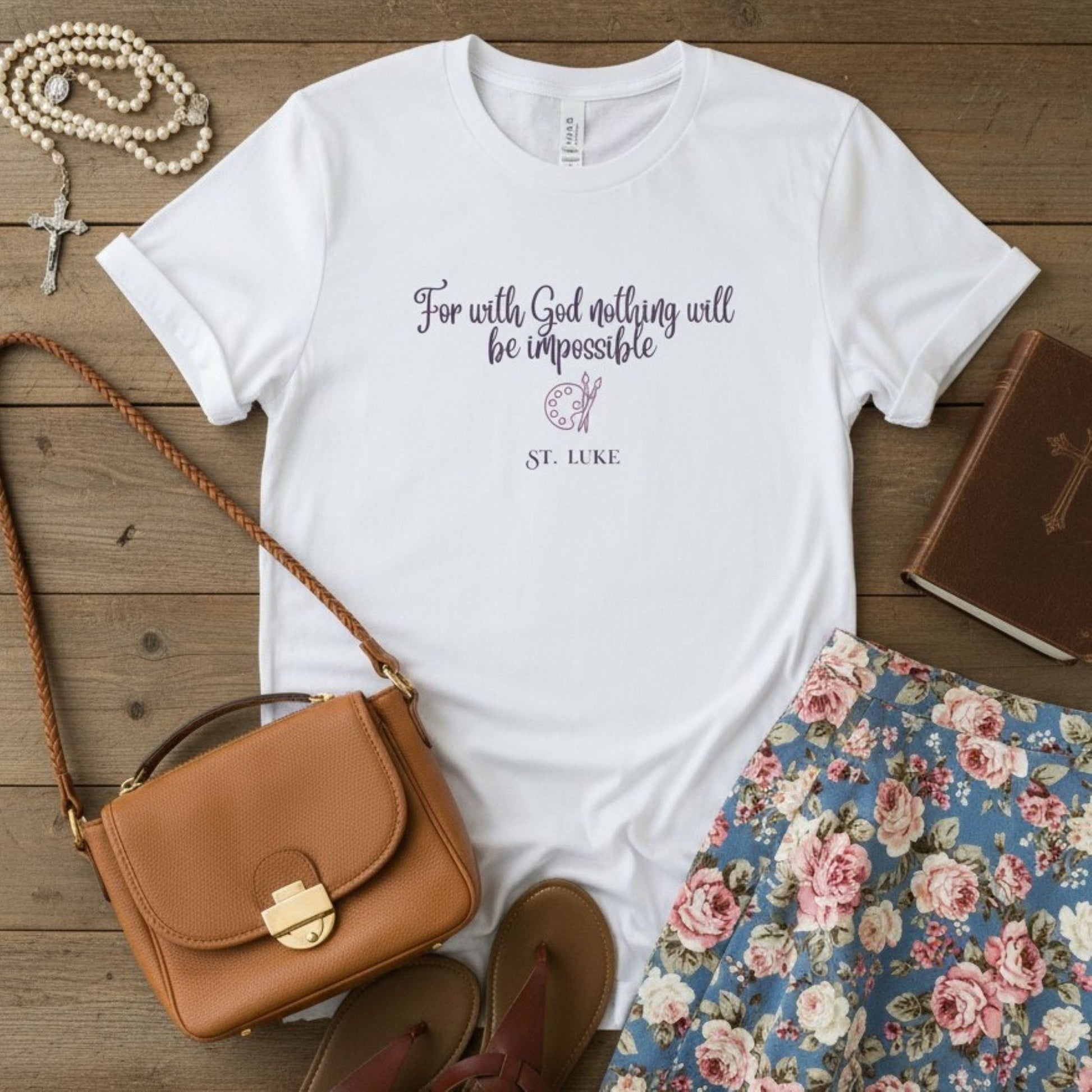 White t-shirt with St. Luke inspirational quote, brown purse, floral skirt, and book on wooden surface
