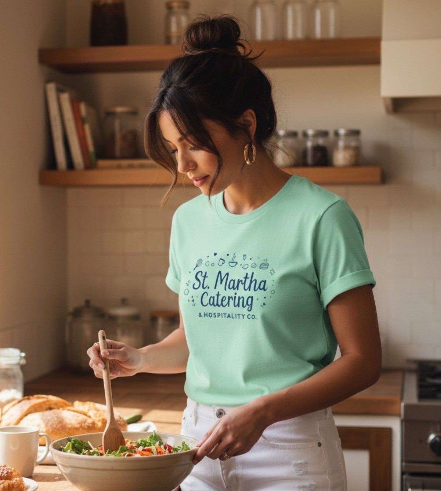 A woman in a kitchen wearing a mint green t-shirt with 'St. Martha Catering & Hospitality Co.