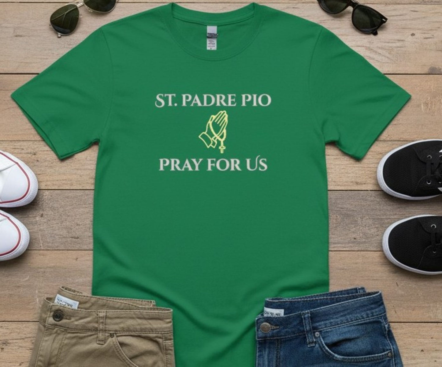 A green short-sleeve T-shirt laid flat on a wooden surface, printed with the words 'ST. PADRE PIO PRAY FOR US' and a small praying hands graphic in white and yellow-green.