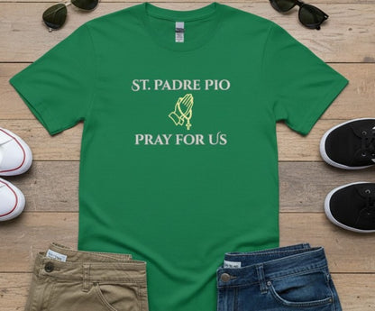 A green short-sleeve T-shirt laid flat on a wooden surface, printed with the words 'ST. PADRE PIO PRAY FOR US' and a small praying hands graphic in white and yellow-green.