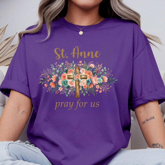 A woman wearing a purple short-sleeve graphic T-shirt with a floral cross design and the words 'St. Anne pray for us' in gold lettering, paired with light blue jeans.