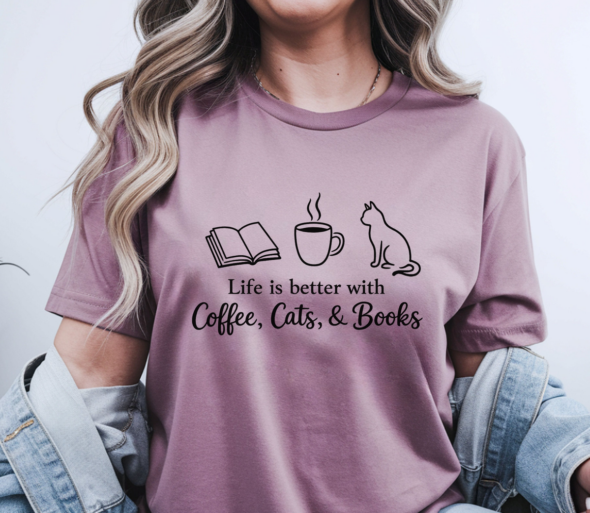 A woman wearing a mauve-purple graphic t-shirt with a black print of an open book, a coffee cup, a cat, and the text 'Life is better with Coffee, Cats, & Books'. She has a light blue denim jacket draped off her shoulders against a plain light gray background.