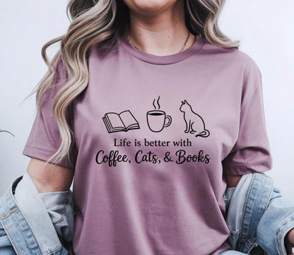 A woman wearing a mauve-purple graphic t-shirt with a black print of an open book, a coffee cup, a cat, and the text 'Life is better with Coffee, Cats, & Books'. She has a light blue denim jacket draped off her shoulders against a plain light gray background.