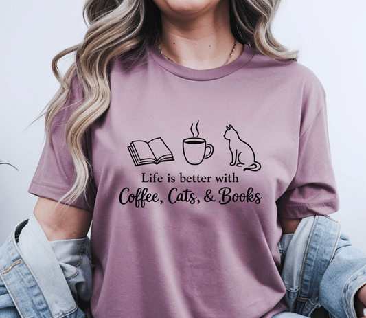 A woman wearing a mauve-purple graphic t-shirt with a black print of an open book, a coffee cup, a cat, and the text 'Life is better with Coffee, Cats, & Books'. She has a light blue denim jacket draped off her shoulders against a plain light gray background.