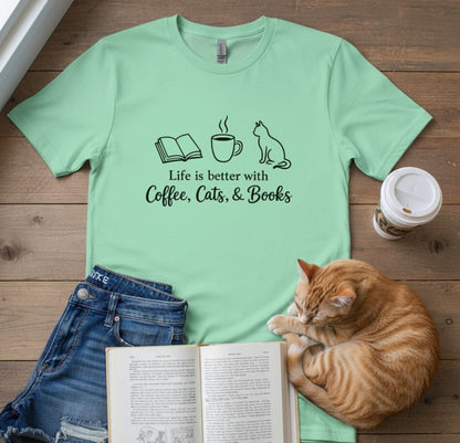 A mint green graphic t-shirt laid flat on a wooden surface, featuring the text 'Life is better with Coffee, Cats, & Books' with simple black line drawings of an open book, a coffee mug, and a cat. The shirt is styled with folded blue jeans, an open book, a sleeping orange tabby cat, and a takeaway coffee cup.