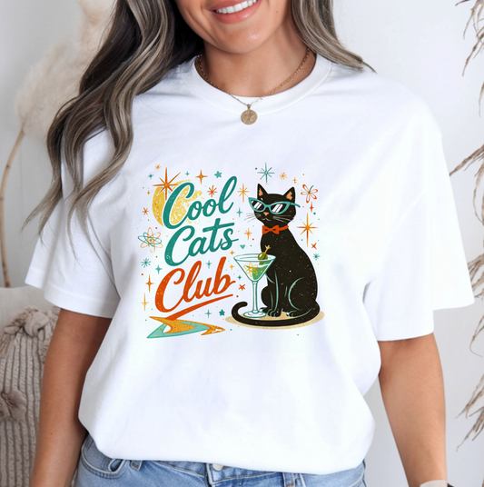 A woman wearing a white short-sleeve graphic T-shirt with a retro-style print of a black cat wearing sunglasses and a bow tie, next to the text 'Cool Cats Club' in teal and orange, paired with blue jeans.