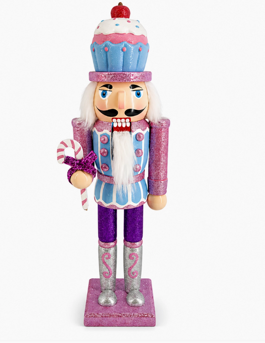 A festive cupcake-themed nutcracker figurine standing on a pink glittery base. The character has a blue and pink cupcake hat, a white beard, a pink and blue outfit with purple accents, silver boots, and holds a pink-and-white striped candy cane against a plain white background.