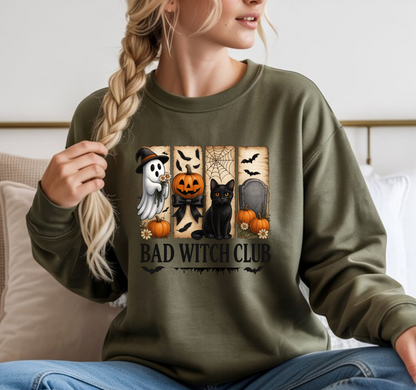 A woman wearing an olive green sweatshirt with a 'Bad Witch Club' Halloween graphic, sitting on a couch.