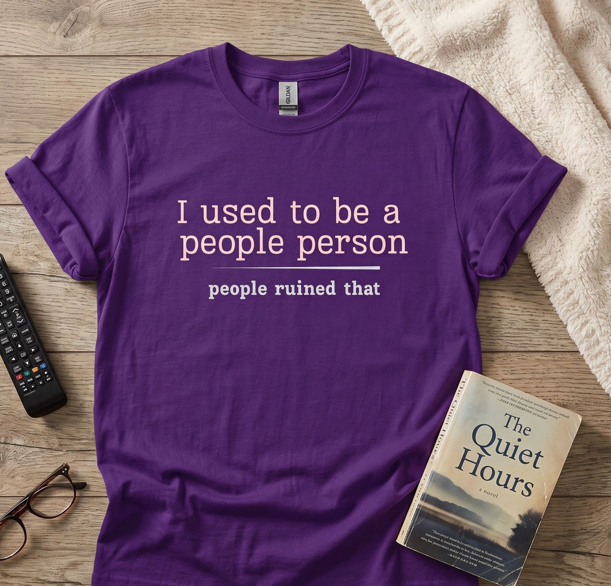 A purple short-sleeve graphic T-shirt laid flat on a wooden surface, with the text 'I used to be a people person people ruined that' printed in white. Surrounding props include a remote control, eyeglasses, a book titled 'The Quiet Hours,' and a cream towel.