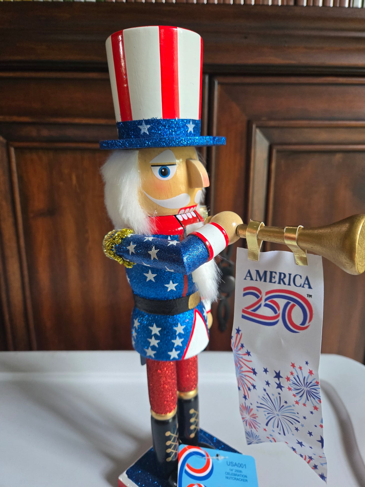 A patriotic nutcracker figurine with a red, white, and blue striped top hat, white beard, and blue star-spangled coat, holding a gold trumpet. A paper tag reading 'AMERICA 2025' is attached, and the background shows a dark wood door and a white surface.