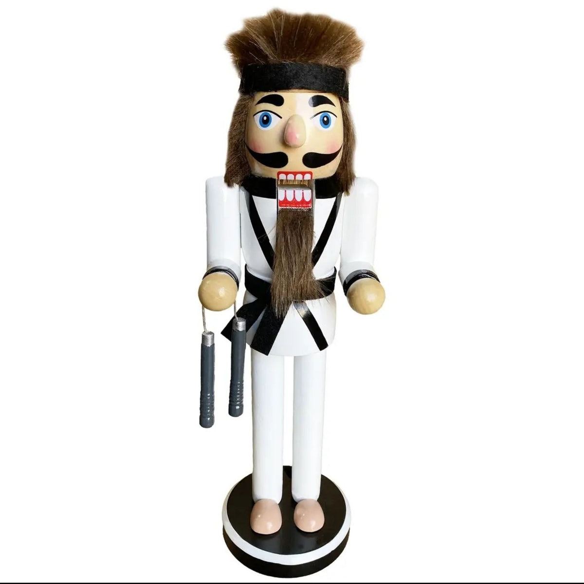 A wooden nutcracker figurine dressed in a white martial arts uniform with black trim, brown hair, and a long brown beard, holding nunchucks and standing on a round base against a white background.