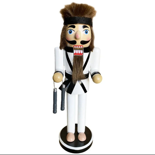 A wooden nutcracker figurine dressed in a white martial arts uniform with black trim, brown hair, and a long brown beard, holding nunchucks and standing on a round base against a white background.