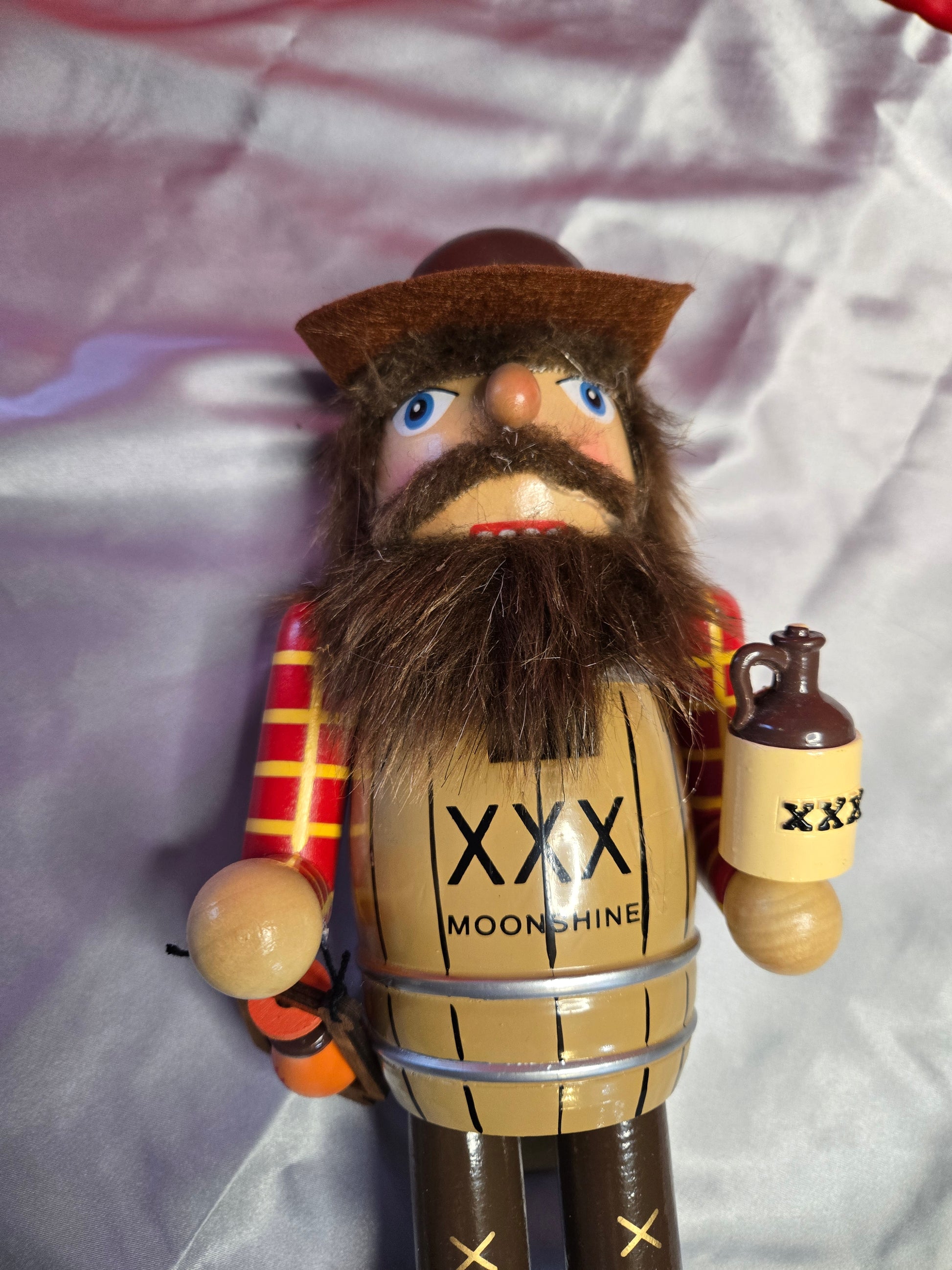 A wooden nutcracker figurine lying on a shiny silver satin fabric background. The nutcracker has a brown hat, a large brown beard, blue eyes, and a tan barrel body with black text reading 'XXX MOONSHINE'.