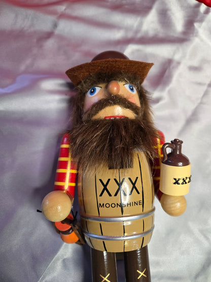 A wooden nutcracker figurine lying on a shiny silver satin fabric background. The nutcracker has a brown hat, a large brown beard, blue eyes, and a tan barrel body with black text reading 'XXX MOONSHINE'.