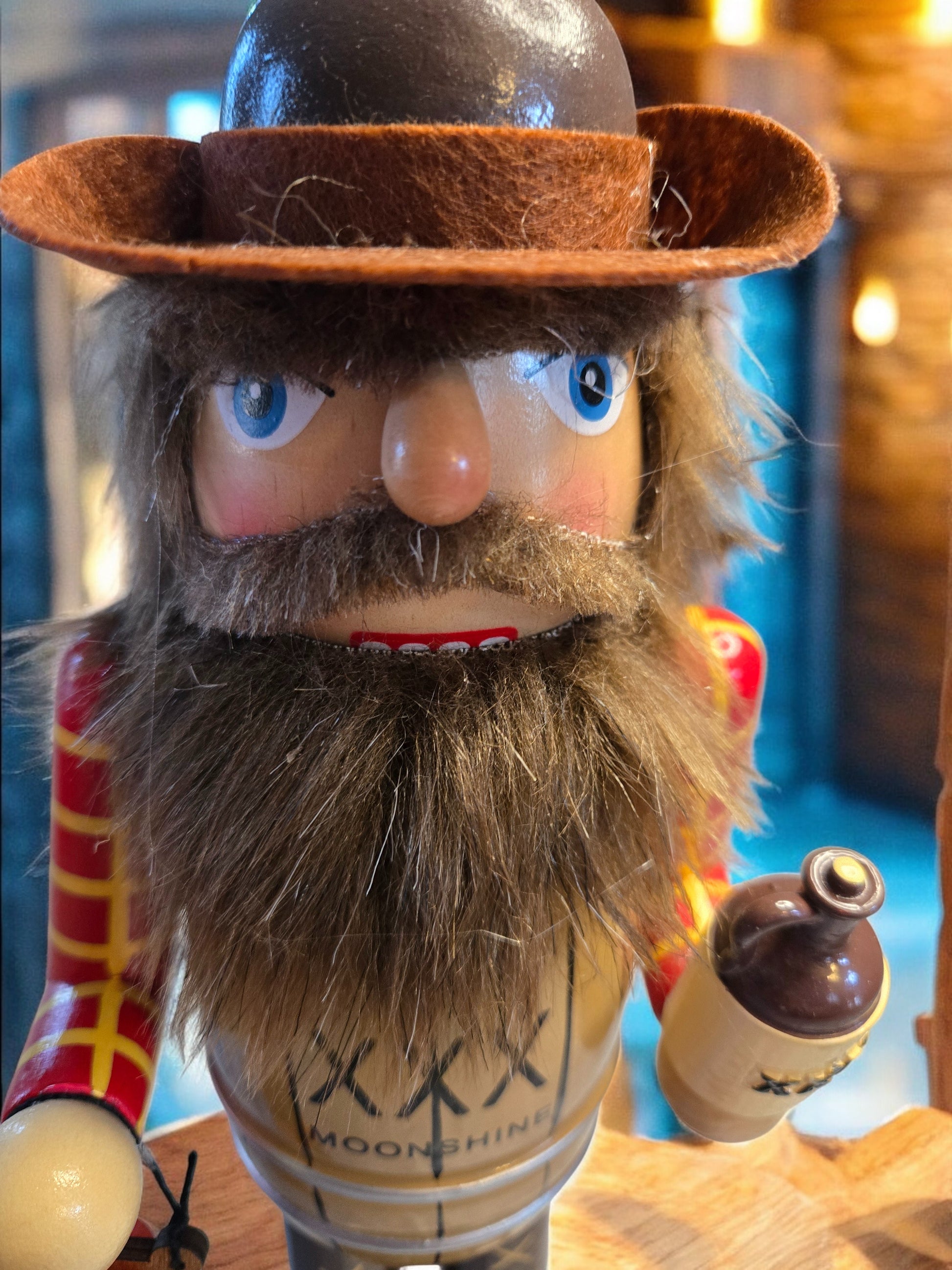 A close-up of a rustic nutcracker-style cowboy figurine with a brown felt hat, large blue eyes, a thick brown beard, and a red-and-yellow striped sleeve, holding a small bottle. The front shows the text 'MOONSHINE XXX'.