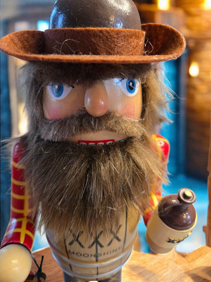 A close-up of a rustic nutcracker-style cowboy figurine with a brown felt hat, large blue eyes, a thick brown beard, and a red-and-yellow striped sleeve, holding a small bottle. The front shows the text 'MOONSHINE XXX'.