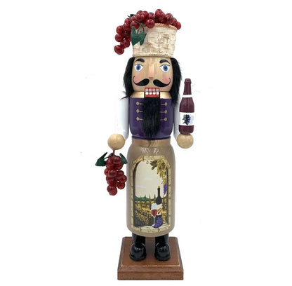A hand-painted wooden nutcracker styled as a wine bottle soldier, featuring a purple vest, black beard, a wine bottle in one hand, and a basket of red grapes on his head, set against a plain white background.