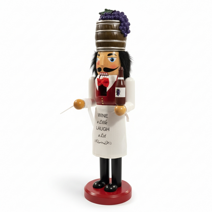 A wine-themed nutcracker figurine standing on a red base, wearing a white apron with the text 'WINE a Little LAUGH a Lot' and holding a wine bottle, with a brown barrel topped with purple grapes on his head.