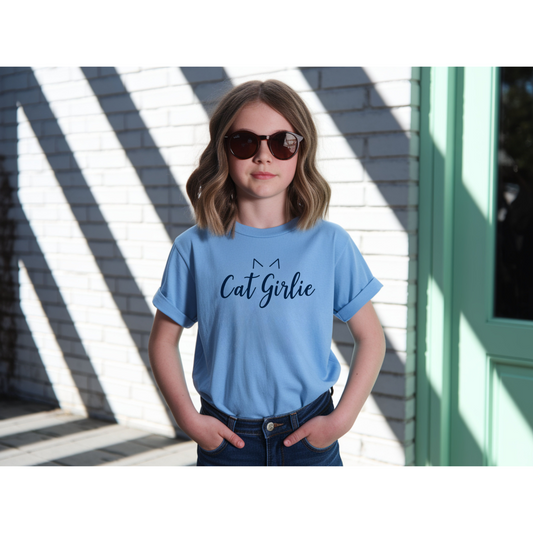 A young girl with wavy brown hair and sunglasses wearing a light blue t-shirt with 'Cat Girlie' printed in navy script, standing outdoors against a white brick wall with diagonal shadows.