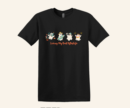 A black short-sleeve crewneck t-shirt featuring a cartoon graphic of four ghosts and the text 'Living My Best Afterlife' in orange, displayed against a solid light beige background.