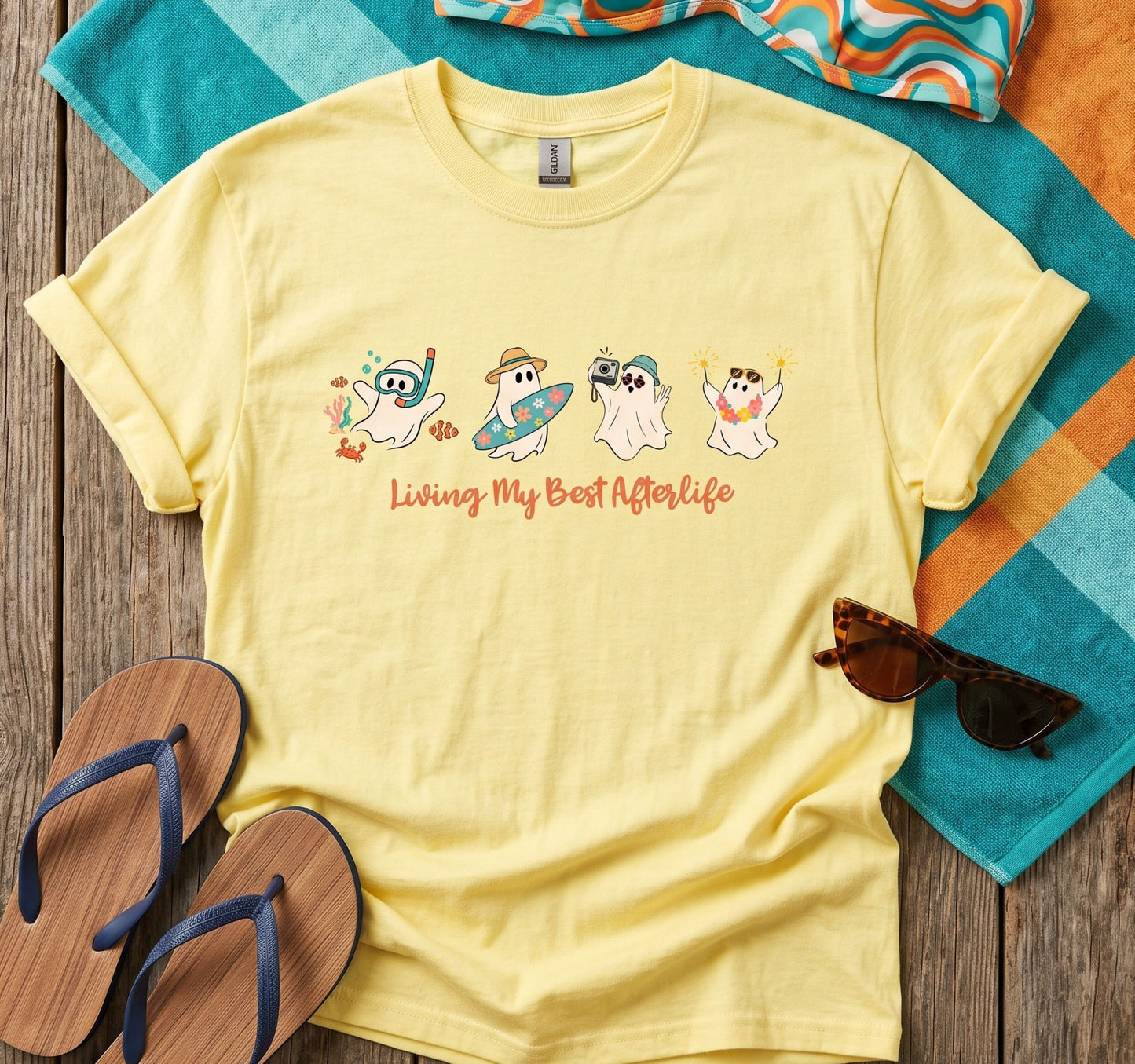 A pale yellow graphic T-shirt laid flat on a wooden surface with beach-themed props including a striped towel, flip-flops, sunglasses, and a patterned bikini top. The shirt has a cartoon chest graphic of four playful ghosts with beach and spa-themed accessories above the text 'Living My Best Afterlife'.