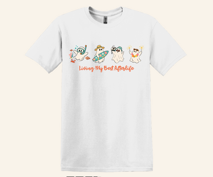 A white short-sleeve crewneck t-shirt featuring a graphic of four cartoon ghosts in different summer poses with the text 'Living My Best Afterlife' in orange script, displayed against a light beige background.