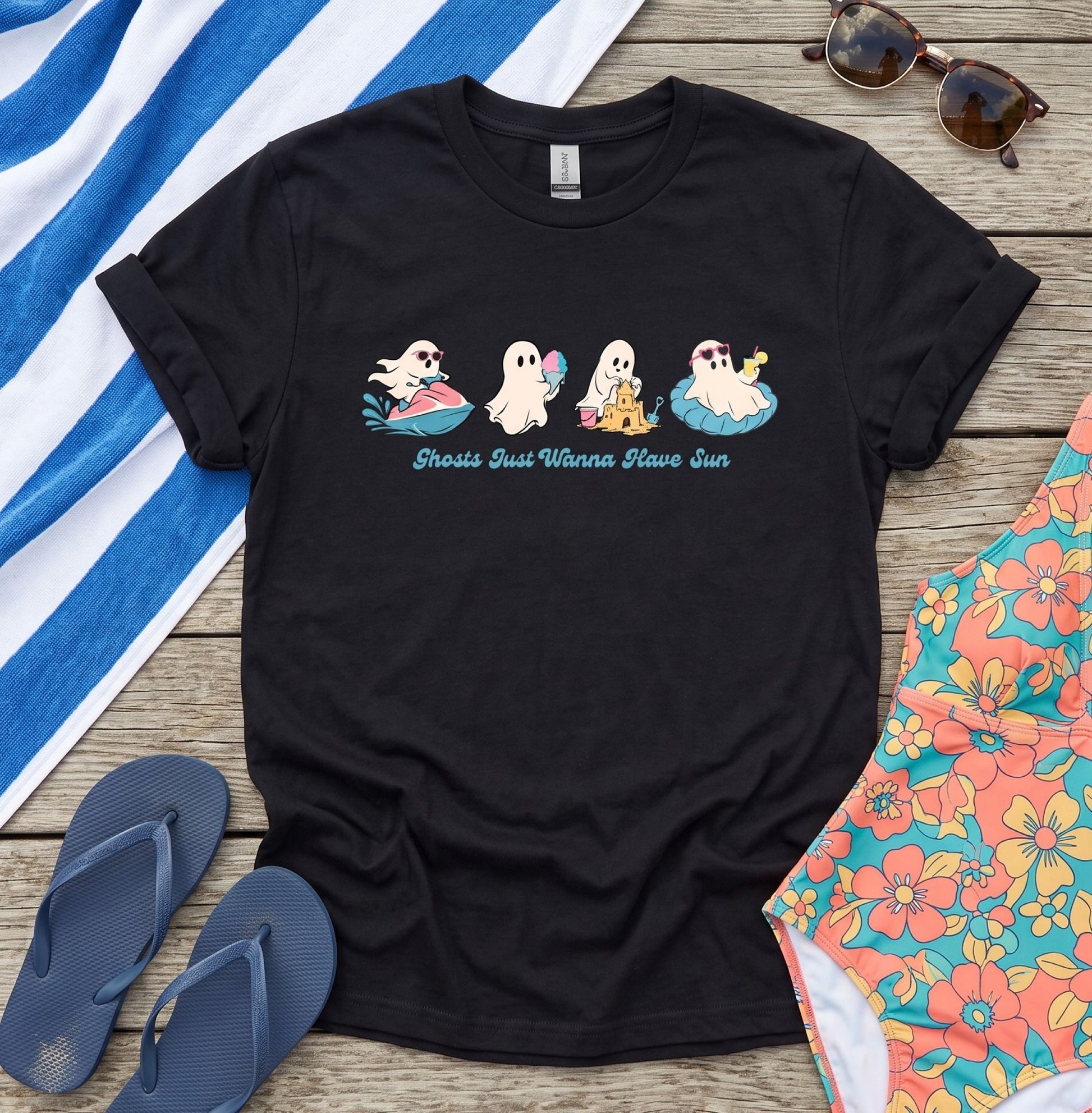 A black graphic t-shirt laid flat on a rustic wooden surface, surrounded by beach-themed props including a blue and white striped towel, sunglasses, flip-flops, and a floral swimsuit. The t-shirt features a graphic of five cartoon ghosts riding surfboards with the text 'Gross Gifs Wanna Blow Sun'.