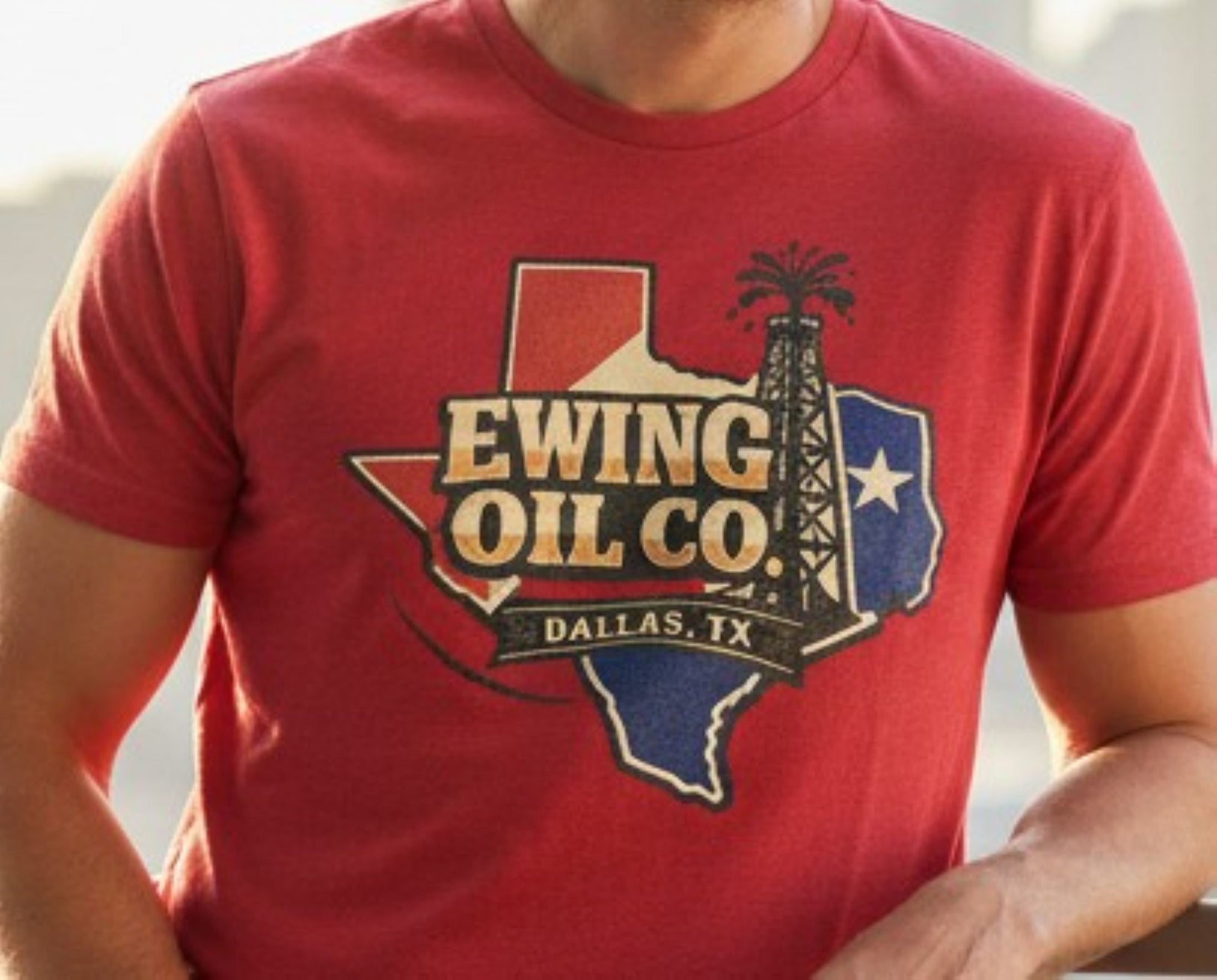 A red short-sleeve crewneck t-shirt featuring a graphic of the state of Texas filled with a red, white, and blue design, overlaid with the text 'EWING OIL CO. DALLAS, TX' and an oil derrick illustration.