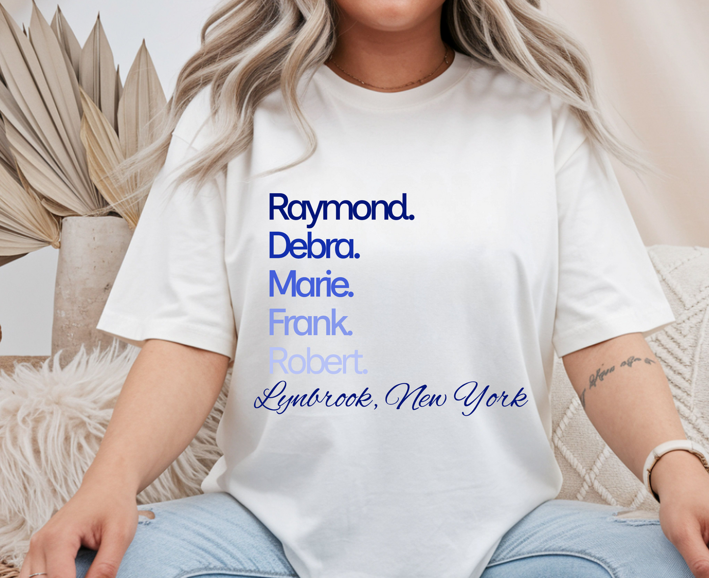 A woman wearing a white short-sleeve graphic T-shirt with stacked names in blue and light blue text reading 'Raymond. Debra.