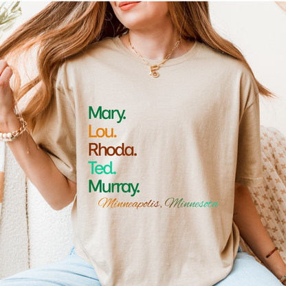 Person wearing a beige t-shirt with names printed on it, sitting on a couch.