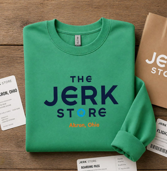 A folded green crewneck sweatshirt with 'THE JERK STORE Akron, Ohio' printed on the front, styled on a wooden surface with themed boarding pass and paper bag props.