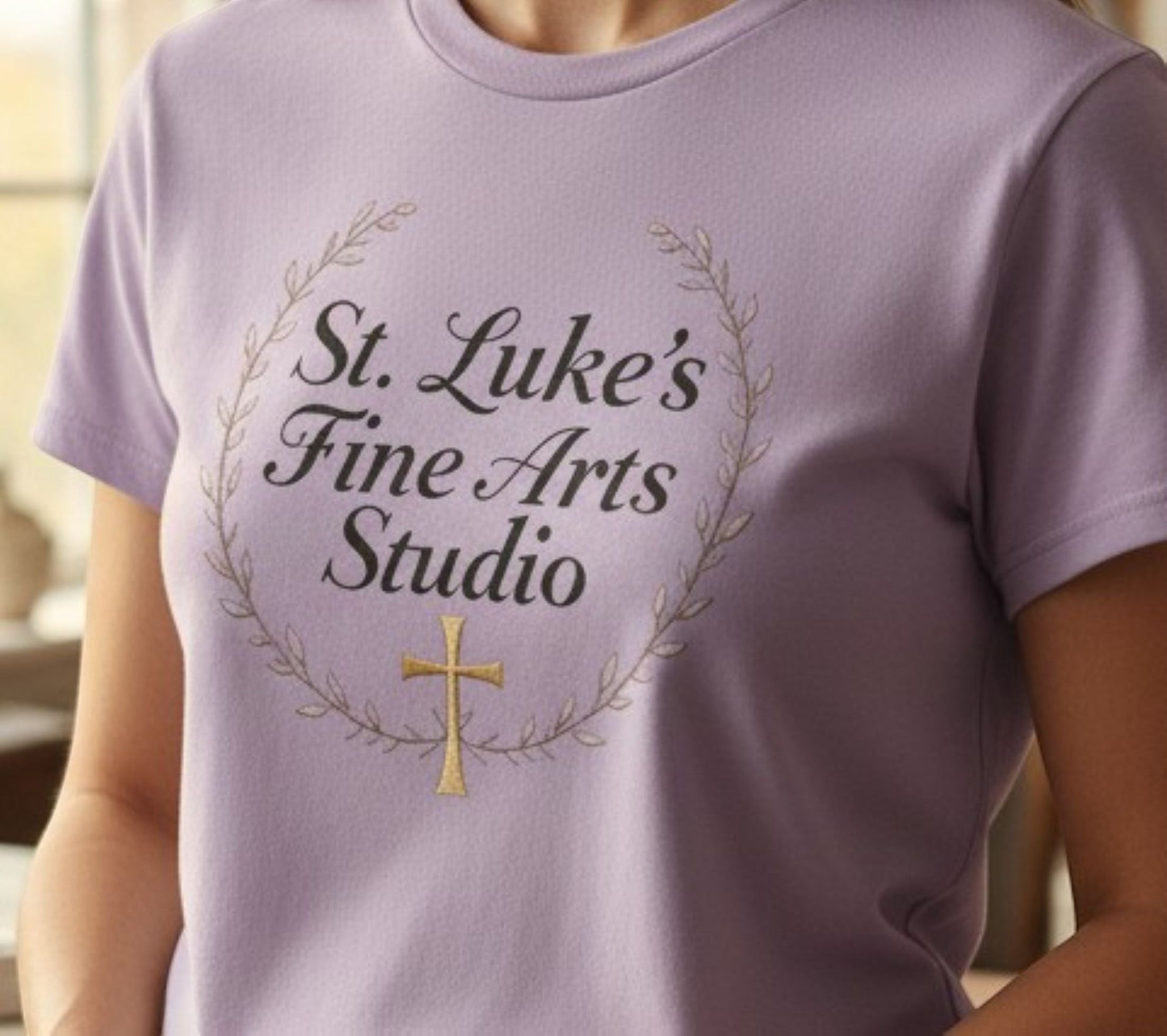 All I Need Designs light purple cotton blend t-shirt with "St. Luke's Fine Arts Studio" and gold cross design.