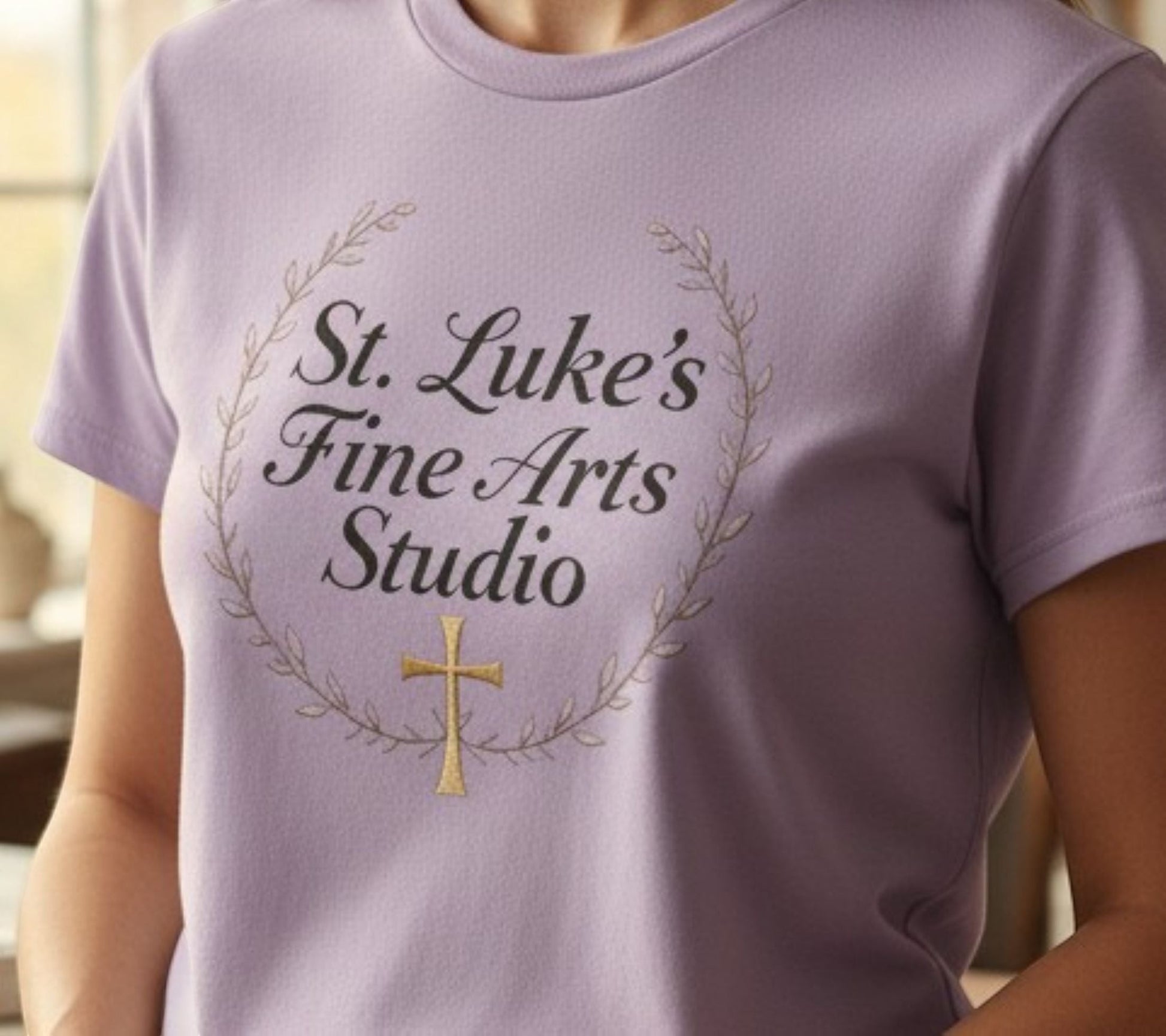 All I Need Designs light purple cotton blend t-shirt with "St. Luke's Fine Arts Studio" and gold cross design.